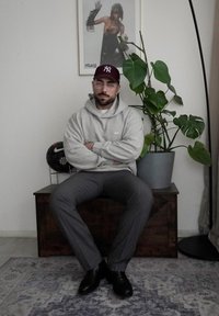 Siva kapuca s logotipom, sivi hlače, crne cipele. Drveni čestak kao sjedište, unutarnja biljka u blizini, i umjetnost na zidu koja prikazuje figuru.