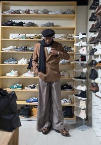 Brauner, doppelt geknöpfter Blazer über einem weißen Hemd, kombiniert mit grauen, weit geschnittenen Hosen und gemusterten Schuhen; schwarzes Barett und Schal. Turnschuhe auf Regalen.