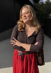 Plava žena s zatvorenim očima stoji na otvorenom, prekriženih ruku, u tamnoj bluzici, crvenoj suknji i smešnom smešnom pojasu, blago se osmehujući na suncu.