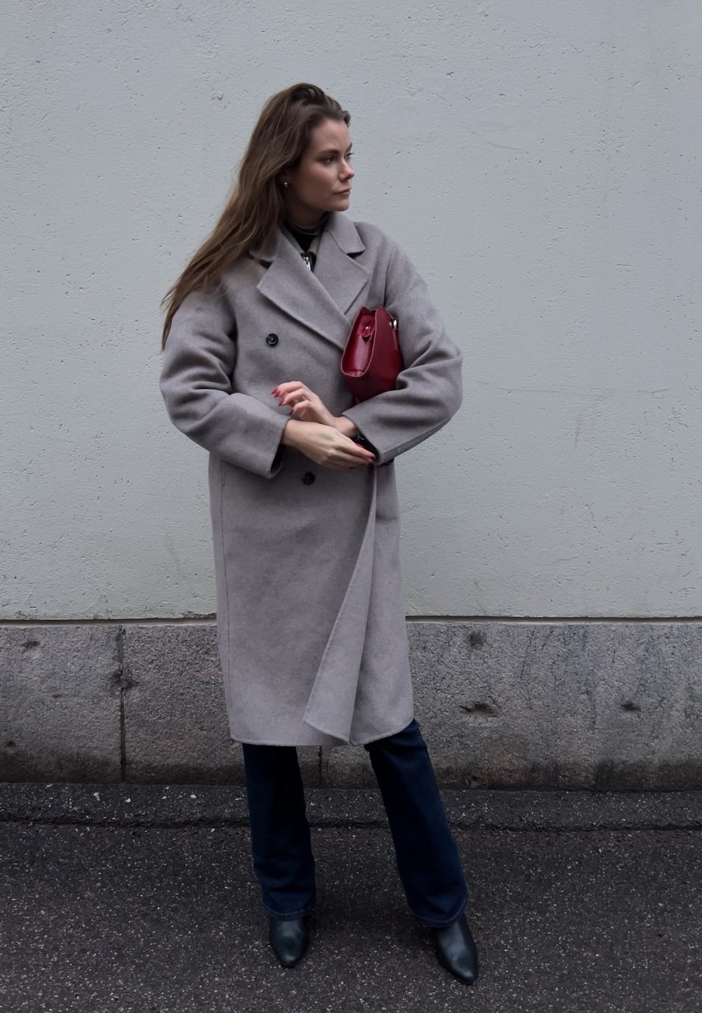 Grigia cappotto di lana con design doppiopetto, indossato sopra pantaloni blu navy e abbinato a una piccola borsa rossa e stivaletti neri alla caviglia.