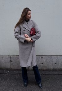 Grigia cappotto di lana con design doppiopetto, indossato sopra pantaloni blu navy e abbinato a una piccola borsa rossa e stivaletti neri alla caviglia.