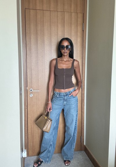 Top marrón ajustado con escote cuadrado, jeans de pierna ancha azul claros, gafas de sol negras, sosteniendo un pequeño bolso marrón, de pie junto a una puerta de madera.