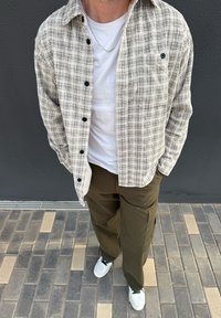 Chemise en flanelle à carreaux beige et noire avec fermeture à boutons, portée par-dessus un T-shirt blanc ; associée à un pantalon cargo vert olive et des baskets blanches.