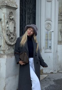 Hall triibuline ülejope, must poolkaelus, valged püksid, hall udune müts ja mustriline pruun käekott. Taustal on rikkalikult kaunistatud arhitektuur.