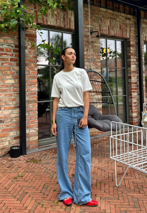 Mujer con camiseta blanca, vaqueros azules de pierna ancha y zapatos rojos está de pie en un patio de ladrillo cerca de una silla colgante y grandes ventanas.