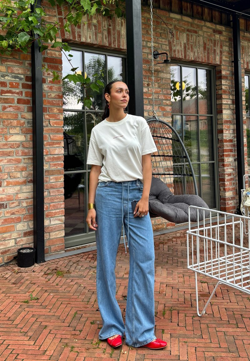 Femme en t-shirt blanc, jean bleu à jambes larges et chaussures rouges, debout sur une terrasse en briques près d'une chaise suspendue et de grandes fenêtres.