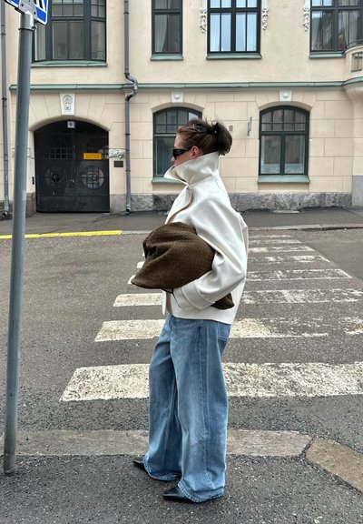Persona con chaqueta blanca, jeans azules holgados y gafas de sol sosteniendo un gran cojín marrón, de pie cerca de un paso de peatones en una calle de la ciudad.