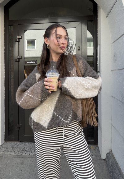 Joven mujer con un suéter marrón estampado y pantalones a rayas sosteniendo un café helado, parada frente a una puerta negra con un bolso de hombro con flecos.