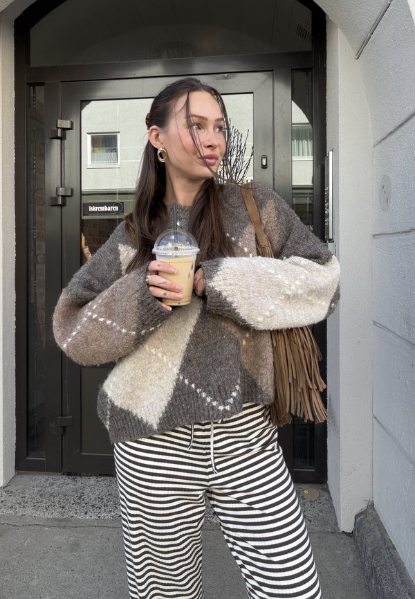 Jeune femme en pull marron à motifs et pantalon rayé tenant un café glacé, debout devant une porte noire avec un sac à franges à l'épaule.