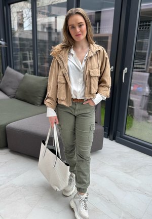 Chaqueta corta beige sobre una camiseta blanca, pantalones cargo verdes y bolsa de mano blanca. Zapatillas de color claro. Texturas suaves, diseño casual.