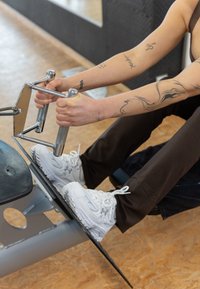 Rameur avec un siège rembourré noir, des poignées en chrome et des poignées texturées. Une personne portant des chaussures de sport blanches est assise, tenant les poignées.