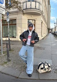 Veste courte en cuir noir, sweat à capuche gris avec logo rouge, jean bleu clair à jambes larges, chaussures noires et sac Puma blanc avec accents noirs.