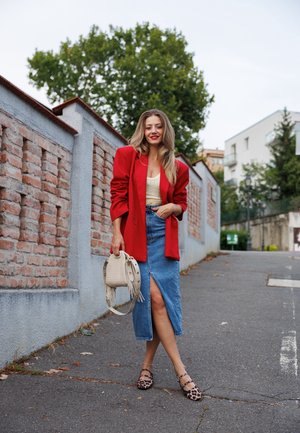 Blazer rojo de gran tamaño, top amarillo claro, falda de mezclilla con abertura en el muslo, zapatos planos de leopardo y bolso beige con detalles de flecos. Entorno urbano.