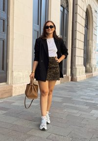 Μαύρο σακάκι πάνω από λευκό μπλουζάκι, φούστα με λεοπάρ μοτίβο, λευκά αθλητικά παπούτσια και καφέ τσάντα. Στηρίζει το σώμα της σε πέτρινο δάπεδο με φόντο κτίριο.