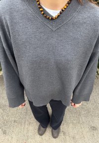 Maglione grigio con scollo a V, collo e polsini a costine, abbinato a pantaloni scuri e una collana di perle con pietre marroni e gialle.