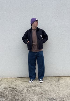 Black jacket over a brown shirt, wide blue jeans, and black and white sneakers. Purple beanie with a white patch, standing against a light wall.