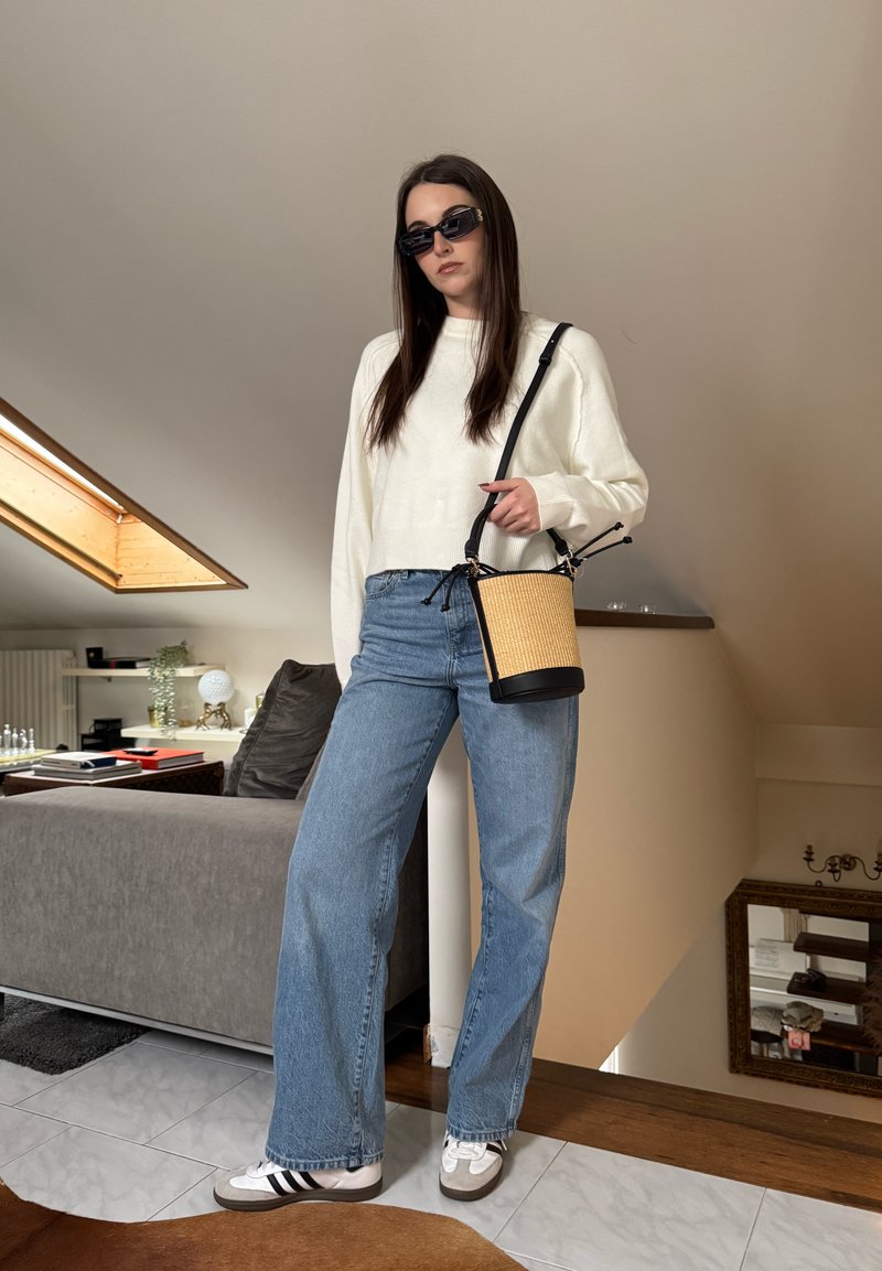 Femme portant un pull blanc, un jean bleu, des lunettes de soleil, des baskets blanches, et tenant un sac seau beige, debout à l'intérieur près d'un canapé et d'une fenêtre de toit.