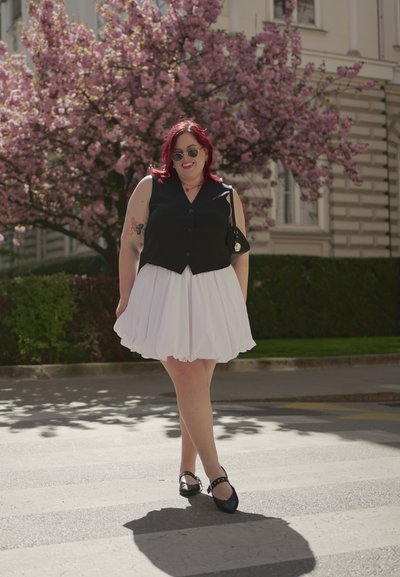 Chaleco negro sin mangas, falda blanca acampanada, zapatos planos negros con tachuelas. Cabello rojo, gafas de sol, de pie en un paso de peatones con árboles en flor rosa.