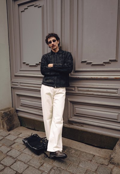 Hombre con cabello rizado y gafas de sol, que lleva una chaqueta negra peluda y pantalones blancos, apoyado en una puerta de madera gris ornamentada con una bolsa negra sobre adoquines.