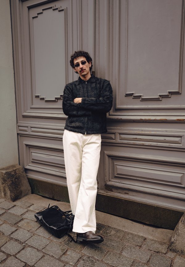 Homme aux cheveux bouclés et lunettes de soleil portant une veste noire duveteuse et un pantalon blanc, appuyé contre une porte en bois grise ornée avec un sac noir sur un pavé.