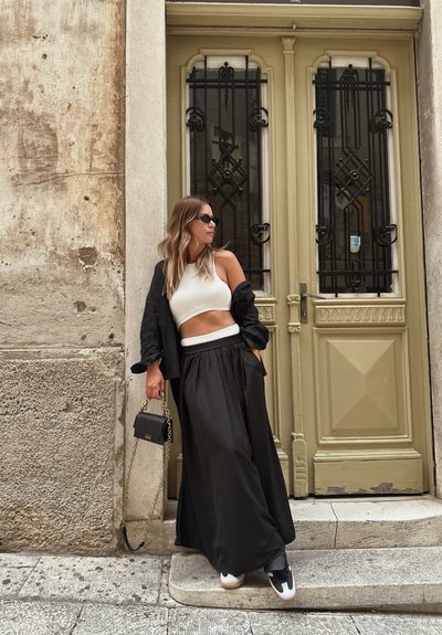 Falda maxi semitransparente negra, top corto blanco, chaqueta negra en capas, bolso pequeño con strap de cadena, zapatillas modernas. Puerta histórica de fondo.
