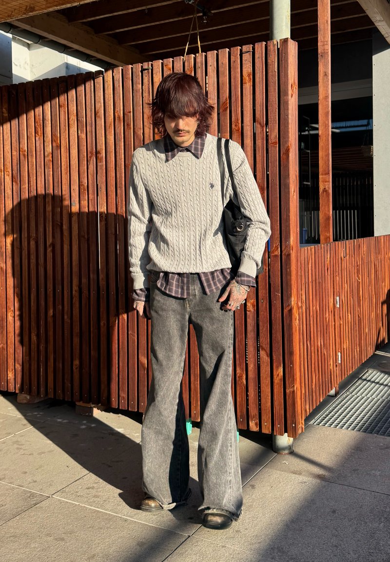 Giovane con capelli alla spalla che indossa un maglione grigio a trecce sopra una camicia a quadri, pantaloni grigi larghi e porta una borsa nera, in piedi vicino a una recinzione di legno.