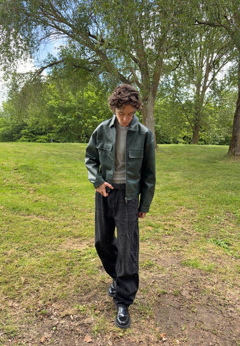 Jeune personne portant une veste vert foncé et un pantalon noir marchant sur l'herbe dans un parc avec des arbres en arrière-plan.