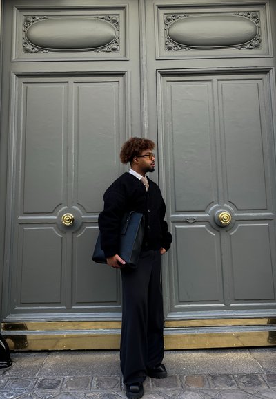 Suéter negro de gran tamaño con botones, camisa blanca y pantalones negros. Sosteniendo un bolso negro. Puertas grises con herrajes dorados ornamentados al fondo.