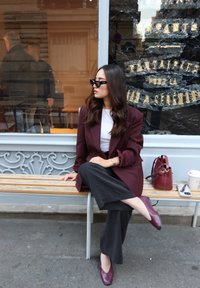 Blazer color melanzana sopra una maglietta bianca, pantaloni larghi neri e scarpe bordeaux. Accessori includono una borsa e occhiali da sole. Panchina in legno e metallo.