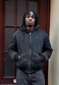 Homme aux dreadlocks portant une veste à capuche noire et un jean, les mains dans les poches, debout devant une porte rouge foncé avec des panneaux de verre.