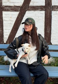 Sort læderjakke, grå hættetrøje og camouflagekasket båret af en person, der holder en lille hvid hund på en blå bænk mod en rustik baggrund.