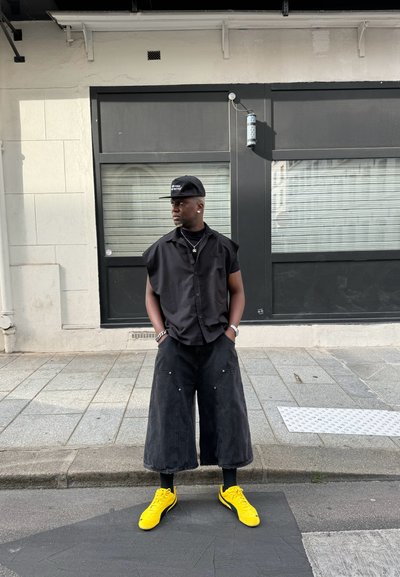 Camisa negra de manga corta, pantalones cortos de mezclilla anchos negros, zapatillas amarillas y una gorra negra con texto blanco. Joyería de plata visible.