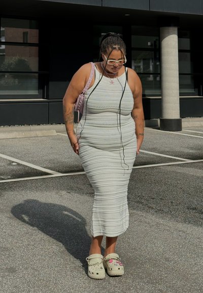 Vestido de punto blanco con rayas horizontales, forma ajustada y escote en forma de barco. Se lleva con zuecos de color claro que presentan decoraciones juguetonas.