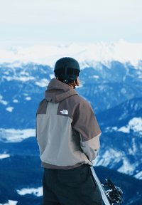 Personne portant un casque de ski et une veste, tenant un snowboard, faisant face à une chaîne de montagnes enneigée sous un ciel clair.