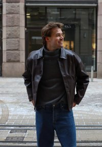 Casaco de pele castanho escuro com fecho de botões, usado sobre um suéter de gola alta cinza e jeans de ganga azul, com uma textura suave e um ajuste descontraído.