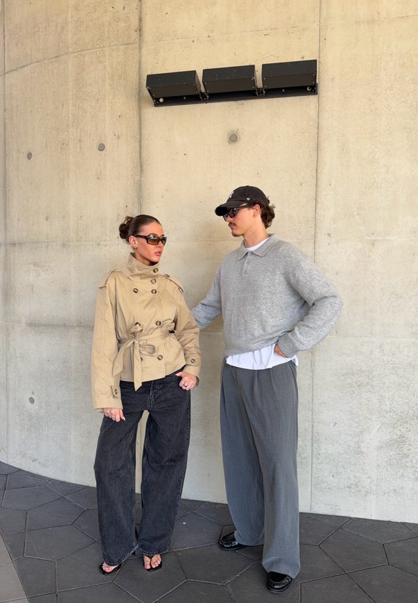 Deux adultes portant des lunettes de soleil se tiennent contre un mur en béton, l'un en veste beige et jean noir, l'autre en pull gris et pantalon ample.