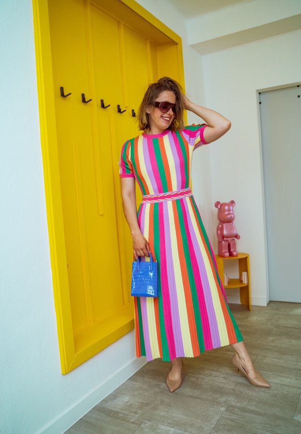 Femme en robe rayée colorée et lunettes de soleil tenant un sac à main bleu, debout près de crochets à manteau muraux jaunes et d'une figurine d'ours rose.
