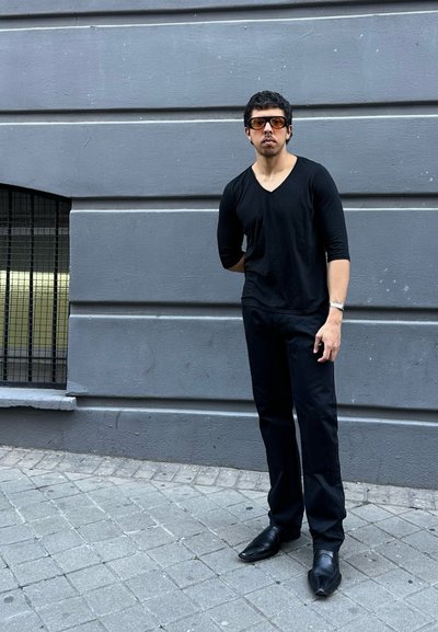 Hombre con camiseta negra de escote en V y pantalones, gafas de sol marrones, de pie en una acera de baldosas grises contra una pared gris con ventana con barrotes.