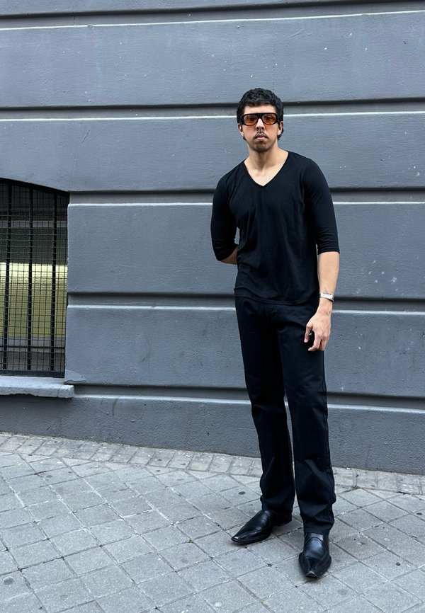 Muškarac u crnoj majici s V-izrezom i hlačama, smeđim sunčanim naočalama, stoji na sivom pločniku uz sivu zidnu površinu s prozorom s rešetkama.