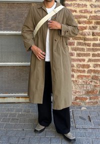 Ελαιόχρωμο παλτό με άνετη γραμμή, διπλό κούμπωμα και πλαϊνές τσέπες. Φορεμένο πάνω από λευκό t-shirt και μαύρα παντελόνια.