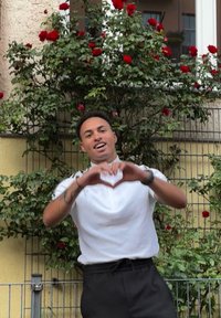 Weißes kurzärmeliges Shirt und schwarze Hose, getragen von einer Person, die mit den Händen eine Herzform macht, umgeben von roten Rosen und grünem Laub.