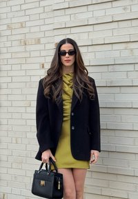 Femme aux longs cheveux ondulés porte des lunettes de soleil noires, un blazer noir, une robe vert olive, et tient un sac à main noir debout devant un mur en briques blanches.