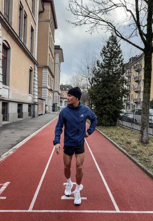 Hombre con chaqueta azul, pantalones cortos negros y gorro de lana, de pie en una pista de atletismo roja en una zona urbana con edificios y árboles sin hojas.