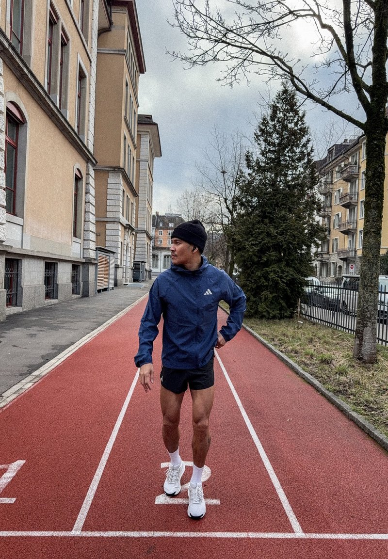 Uomo con giacca blu, pantaloncini neri e berretto in piedi su una pista di atletica rossa in un'area urbana con edifici e alberi senza foglie.