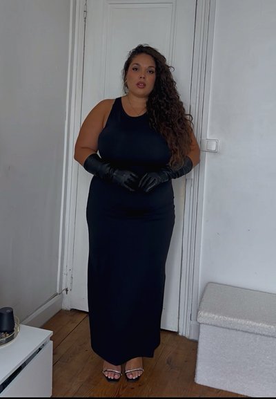 Vestido maxi negro sin mangas, tela suave, forma ajustada. Guantes negros, sandalias de tacón alto con acentos de strass, fondo de suelo de madera.