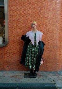 Długa zielona sukienka w kratę z warstwowym różowym kołnierzem i czarnym, oversize'owym płaszczem. Czarne buty na platformie oraz mała różowa torebka z pluszowymi akcentami.