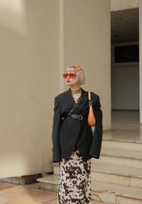 Sort oversized blazer, hvid og brun mønstret midi-nederdel, røde solbriller, lagede guld halskæder, kort pastelfarvet hår og orange taske.