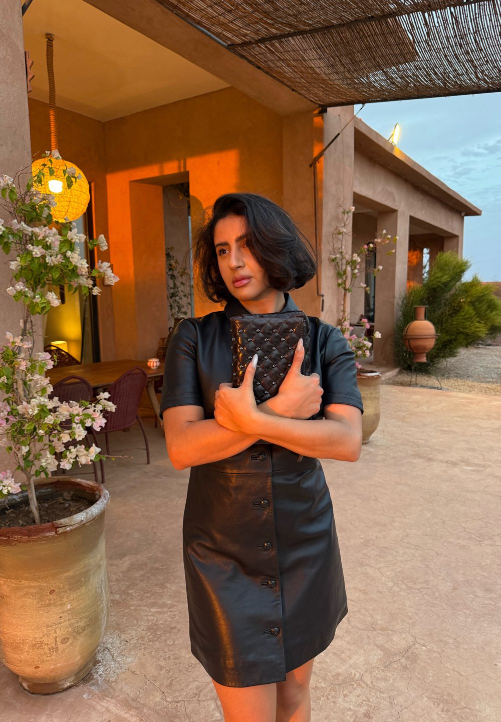 Czarna skórzana sukienka na guziki z krótkimi rękawami, o dopasowanym kroju. Modelka trzyma pikowaną czarną kopertówkę. W tle rośliny i ciepłe oświetlenie.