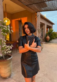 Czarna skórzana sukienka na guziki z krótkimi rękawami, o dopasowanym kroju. Modelka trzyma pikowaną czarną kopertówkę. W tle rośliny i ciepłe oświetlenie.