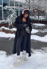 Manteau noir oversize, body noir, chaussettes en maille, bottes à plateforme grises. Porte des écouteurs roses, tient une pochette rayée, sol couvert de neige.